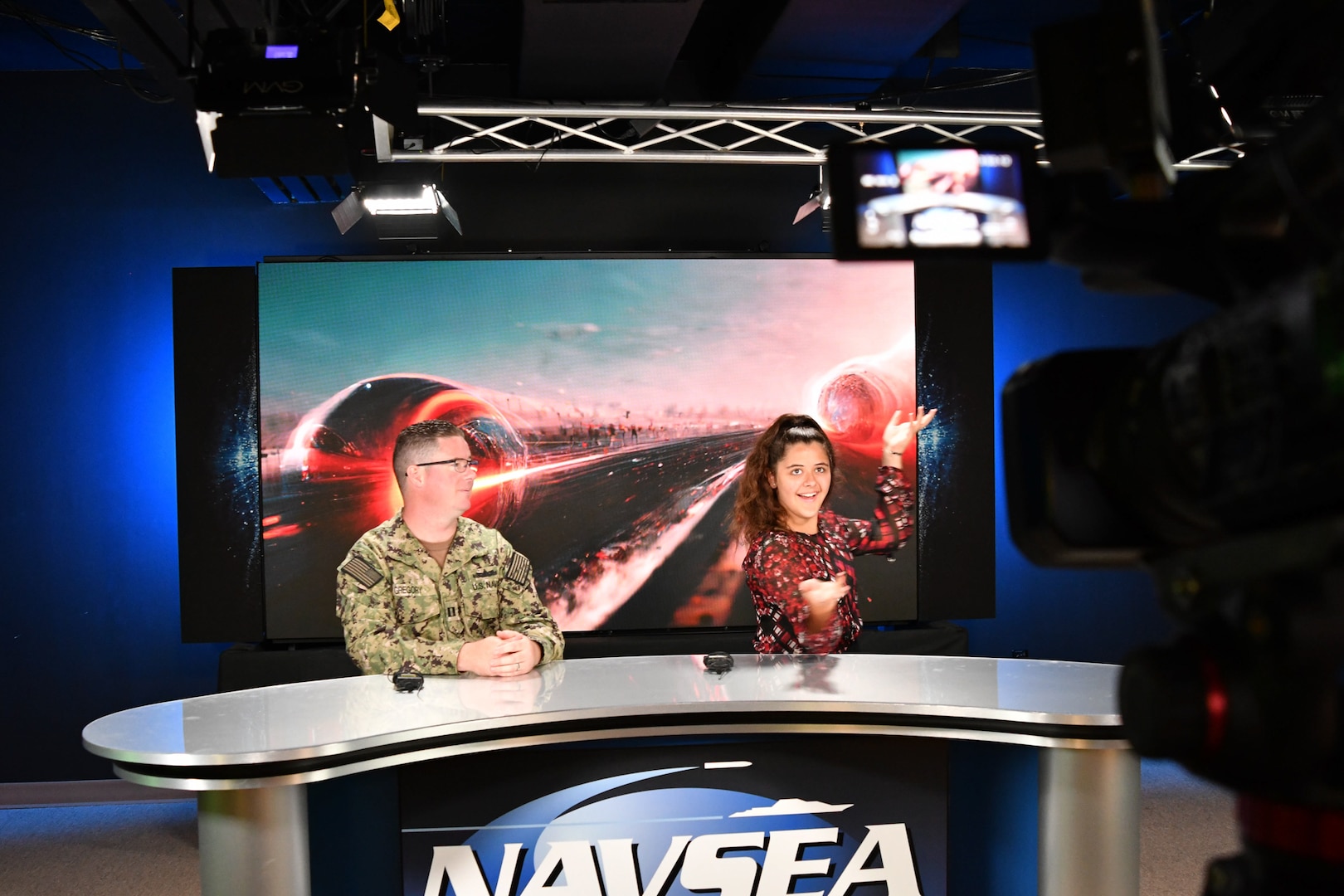 IMAGE: Lt. Marc Gregory and his daughter Mila, 13, joke around behind the broadcast production news desk at the Graphics and Imaging Branch during the Bring Your Child to Work Day event, Aug. 2. The branch provided children in attendance a tour of the professional studio, demonstrations of the camera and recording equipment and the opportunity to appear on screen.