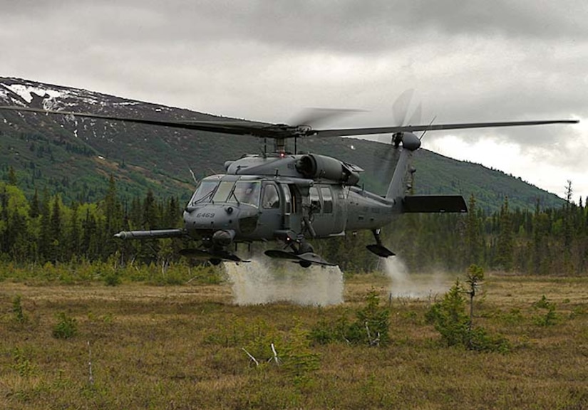 An HH-60G Pave Hawk hovers in a field with mountains in the background.