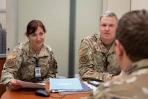 Two military members sit on the opposite end of a table, wearing inspection badges, and looking over papers while speaking to an Airman on the near side of the table.
