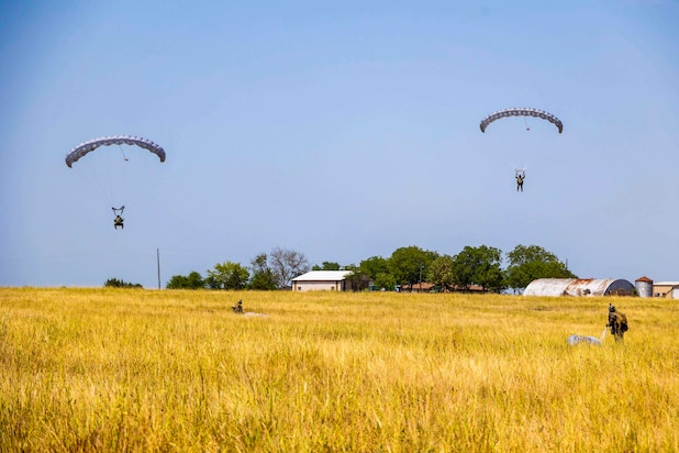 Two Marines descend in the sky wearing parachutes while others walk in a grassy field below.