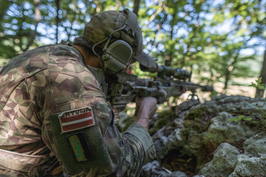 A soldier with a patch that reads “LATVIJA,” which is the native spelling for Latvia, looks down the scope of a rifle in a wooded environment.