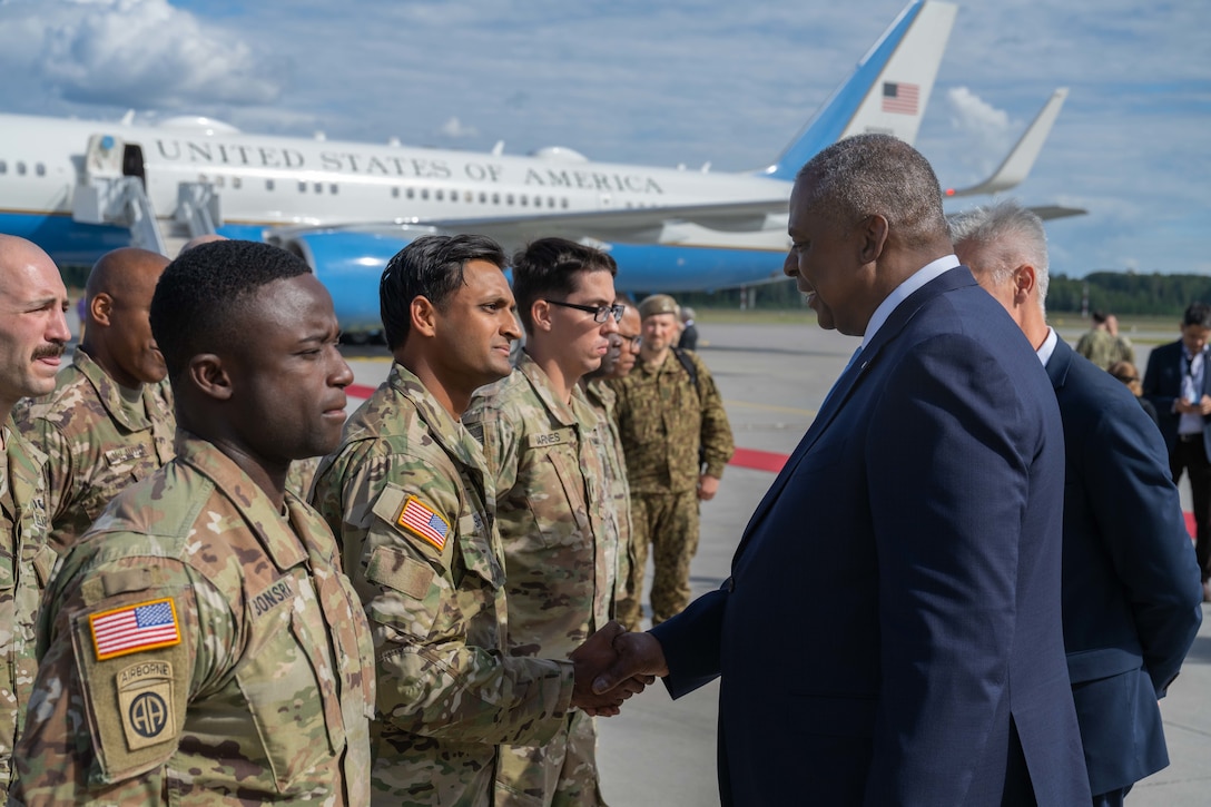 A man in formal business attire shakes hands with a soldier standing in formation in an airfield. A U.S. passenger aircraft can be seen in the background.