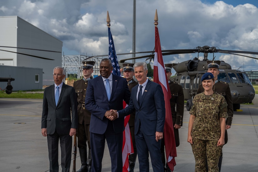 Two men shaking hands with flags, foreign service members and a helicopter in the background.