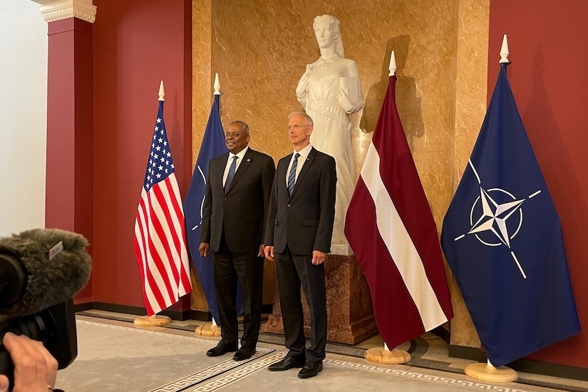 Two men wearing business suits stand shoulder-to-shoulder with flags and a statue in the background.