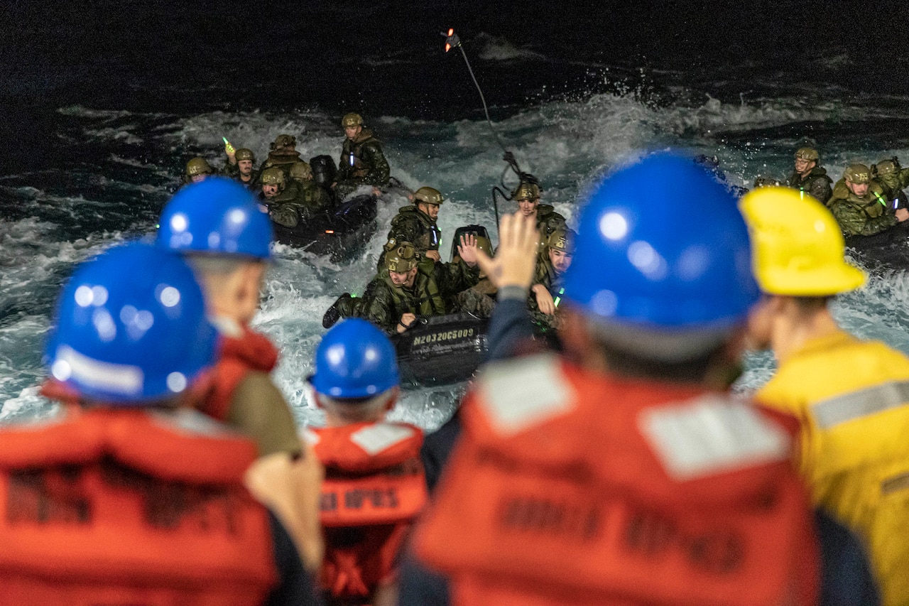 Marines in rubber boats toss lines toward sailors, shown from behind, on a ship at night.