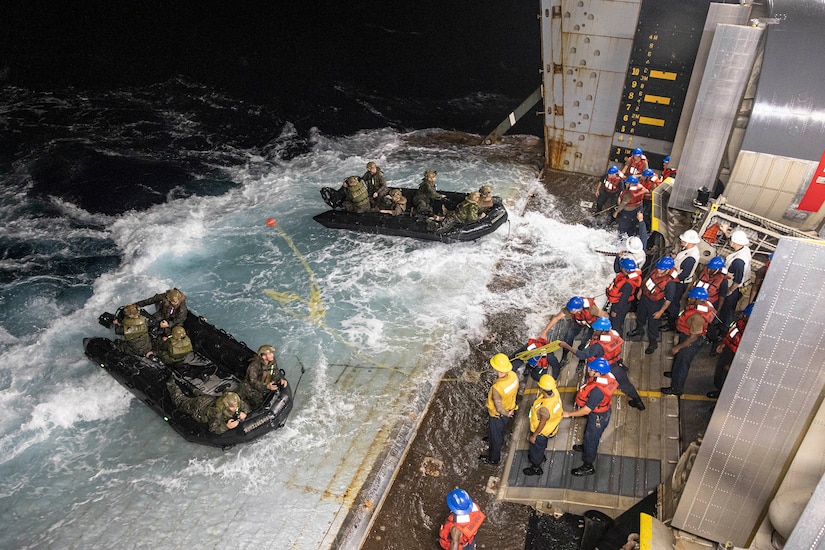 Marines in two rubber boats on a ship's deck throw lines to sailors on the ship.