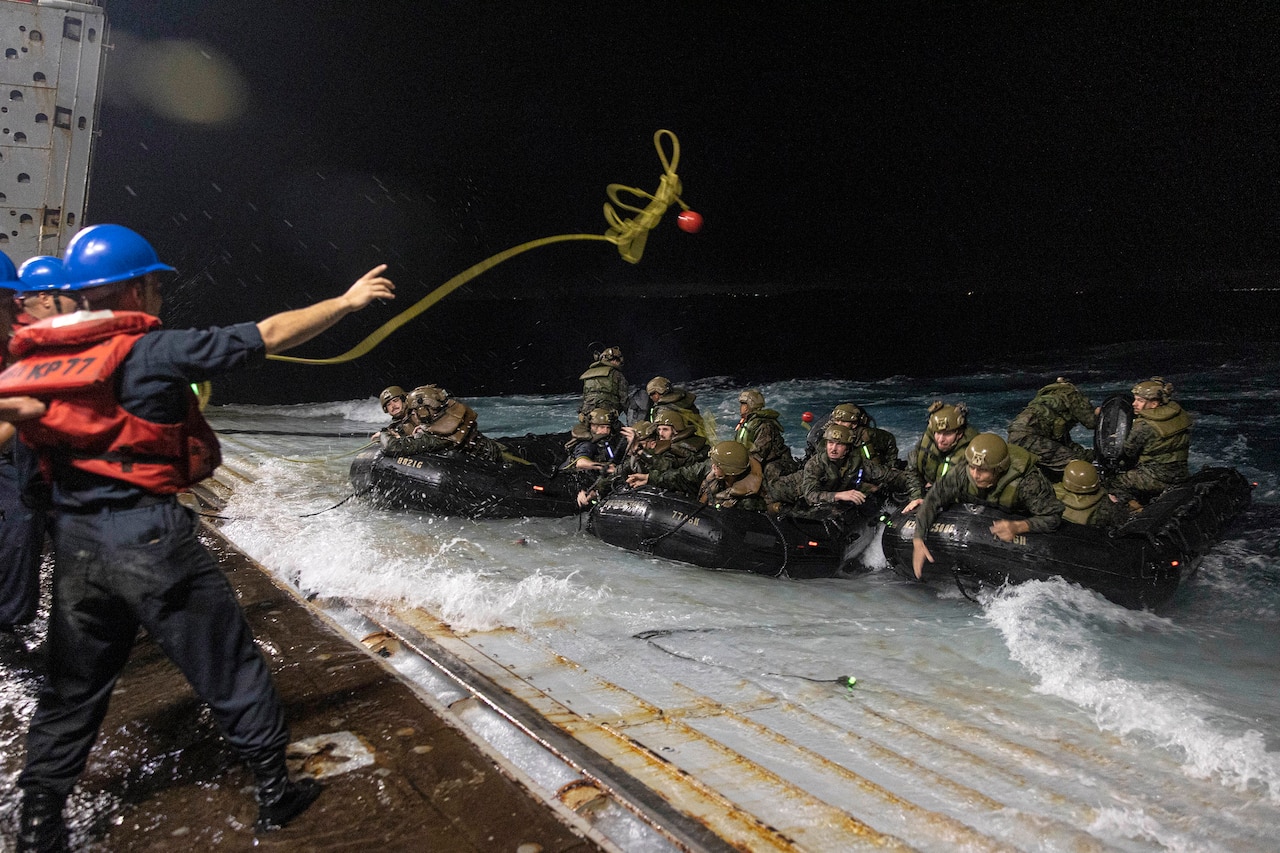 A sailor throws a line from a ship's well deck to Marines in rubber boats.