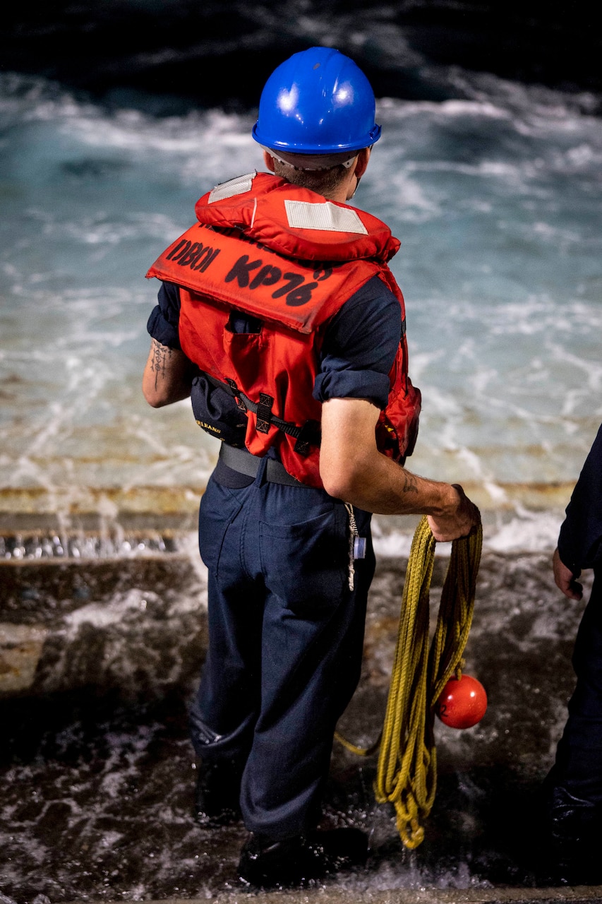 A sailor shown from behind holds a line on a ship's deck.