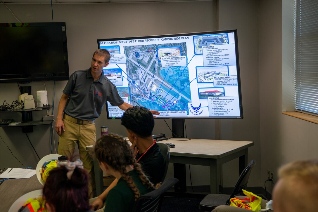 A figure in a grey polo shirt stands in front of a large computer monitor with a map of construction plans