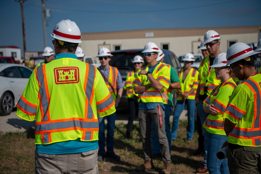 A senior engineer with his back to the camera, wearing a white hard hat with a red stripe and a yellow safety vest with the USACE logo on the back, briefs interns