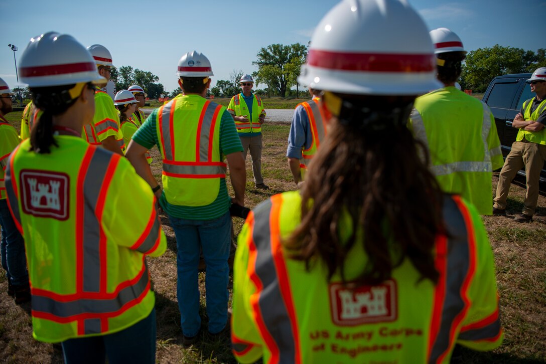 Subjects wearing hard hats and safety vests with the US Army Corps of Engineers logo listen to a figure providing a safety brief