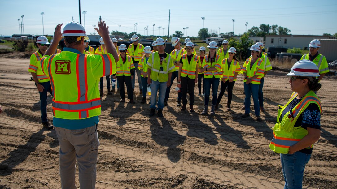 A group of interns wearing hard hats and yellow safety vests listens to a safety and history brief
