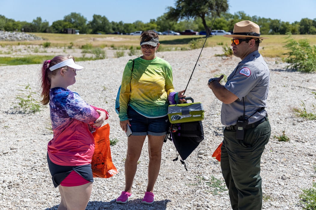 a park ranger in uniform talks to a child and her mother on the shoreline of a lake