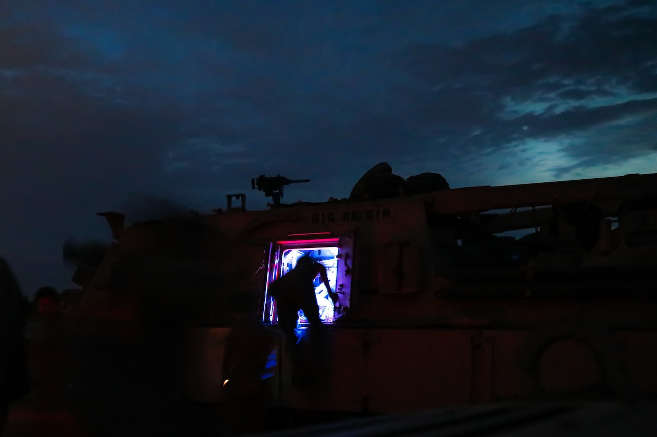 A person leans into the hatch of a military vehicle at night and is silhouetted against a bluish light from inside.