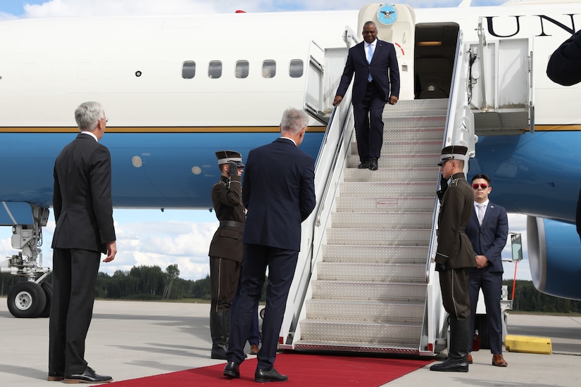 A man in business attire exits an aircraft, descending stairs toward a greeting party and a red carpet.