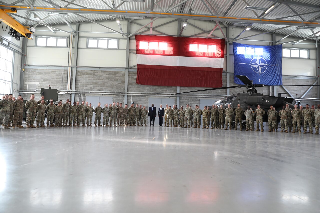 Two men in formal business attire stand at the center of a large formation of service members and pose for a photo in a hangar bay.