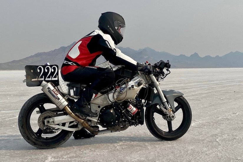 A man wearing a motorcycle suit sits on a racing motorcycle on a salt flat.