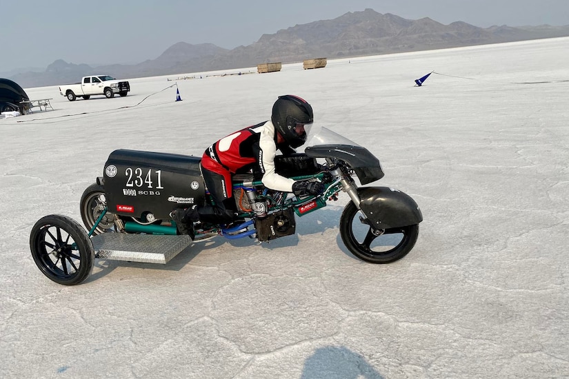 A man wearing a motorcycle suit sits on a three-wheeled racing motorcycle on a salt flat.