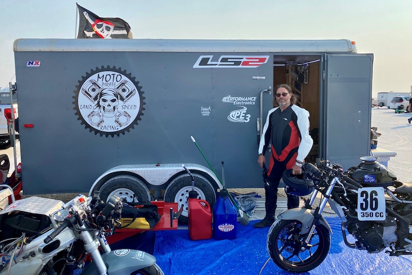 A man wearing a motorcycle suit stands near two racing motorcycles with a small trailer in the background.