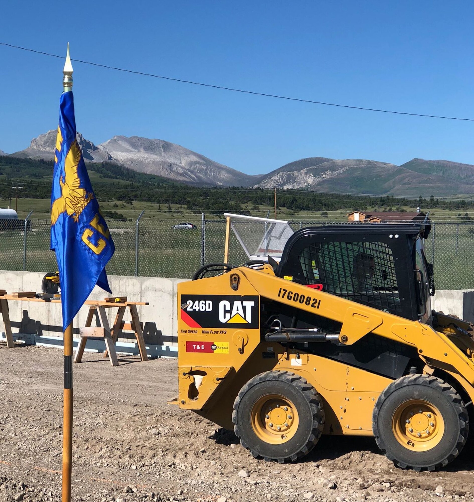 a piece of construction equipment sitting adjacent to a unit guidon outside with a mountain range in the distance.