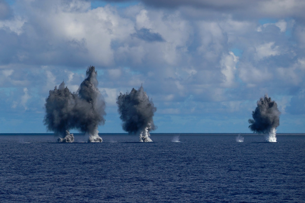 Explosions create grey smoke clouds above water.
