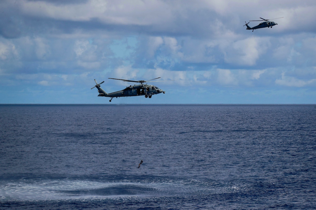 Two helicopters fly over water, with a person hanging from one by a line.