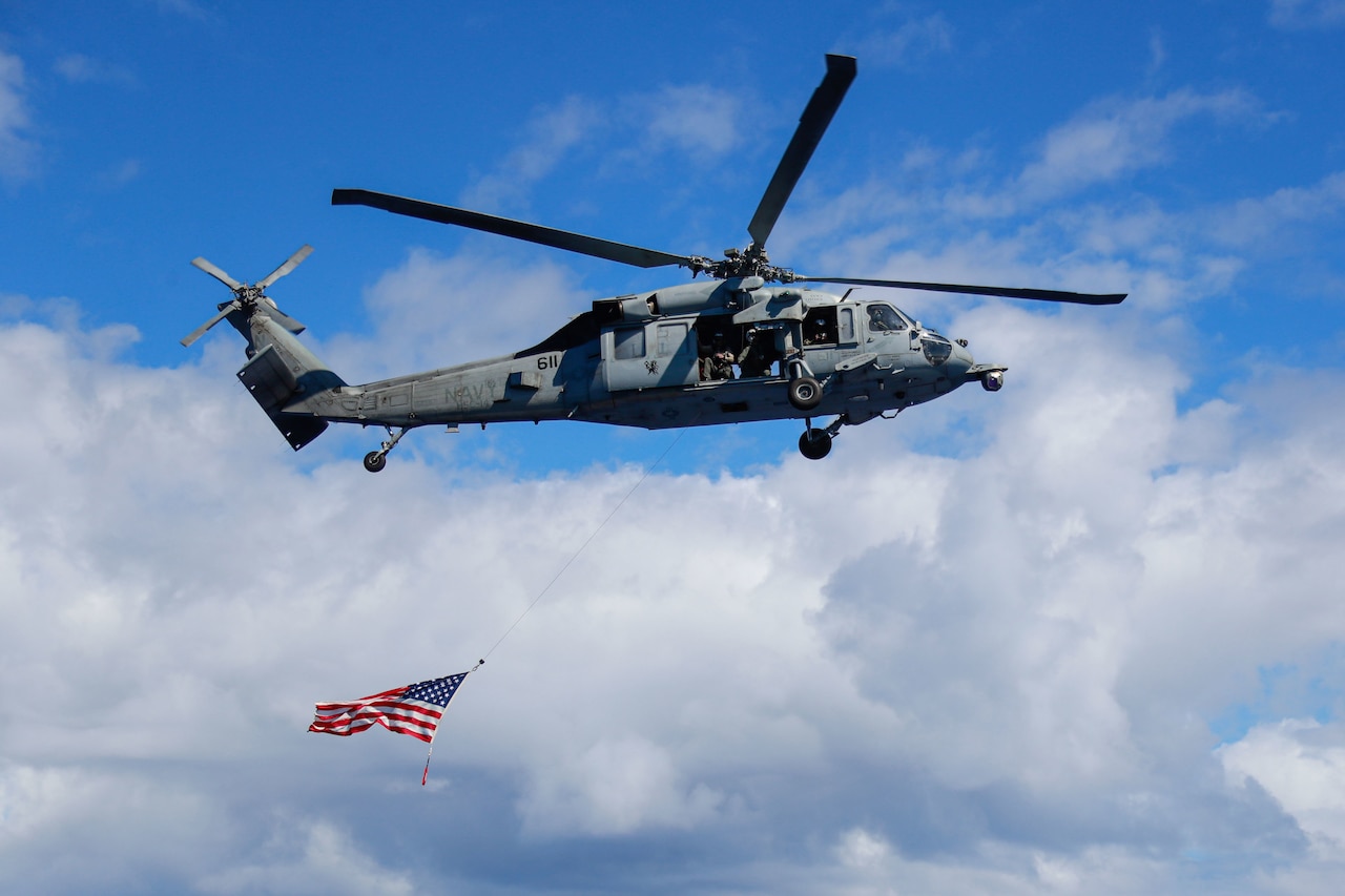 A flag attached by line to a flying helicopter waves beneath it in blue sky.