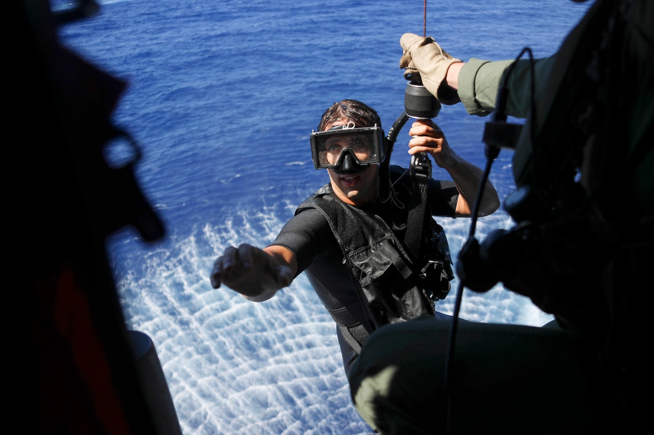 A sailor attached to a hoist reaches toward troops in a helicopter over water.