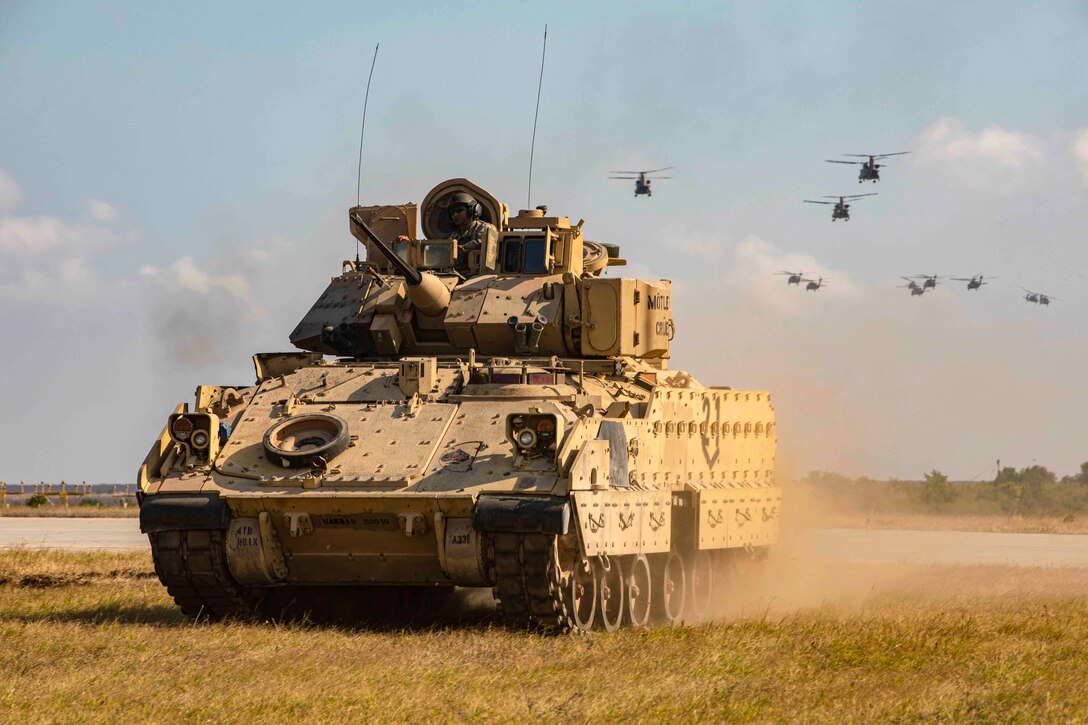 A military vehicle moves across a grassy field as a group of helicopters fly in the background.