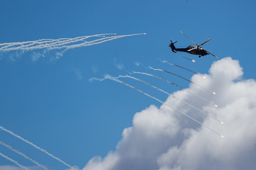 Flares shoot out behind a helicopter in cloud-studded blue sky.