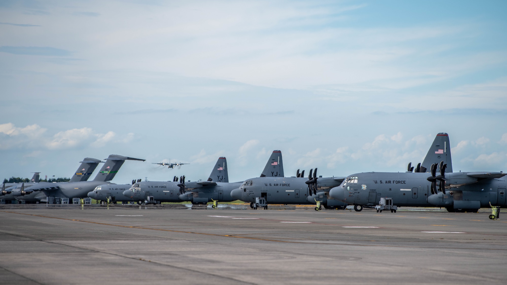 A row of parked C-130J Super Hercules sit on the flight line while a Royal Thai Air Force C-130H Hercules lands on a runway