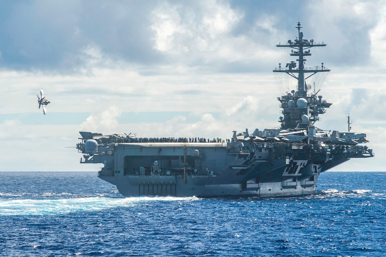 An aircraft flies close to a ship at sea with people on the flight deck.