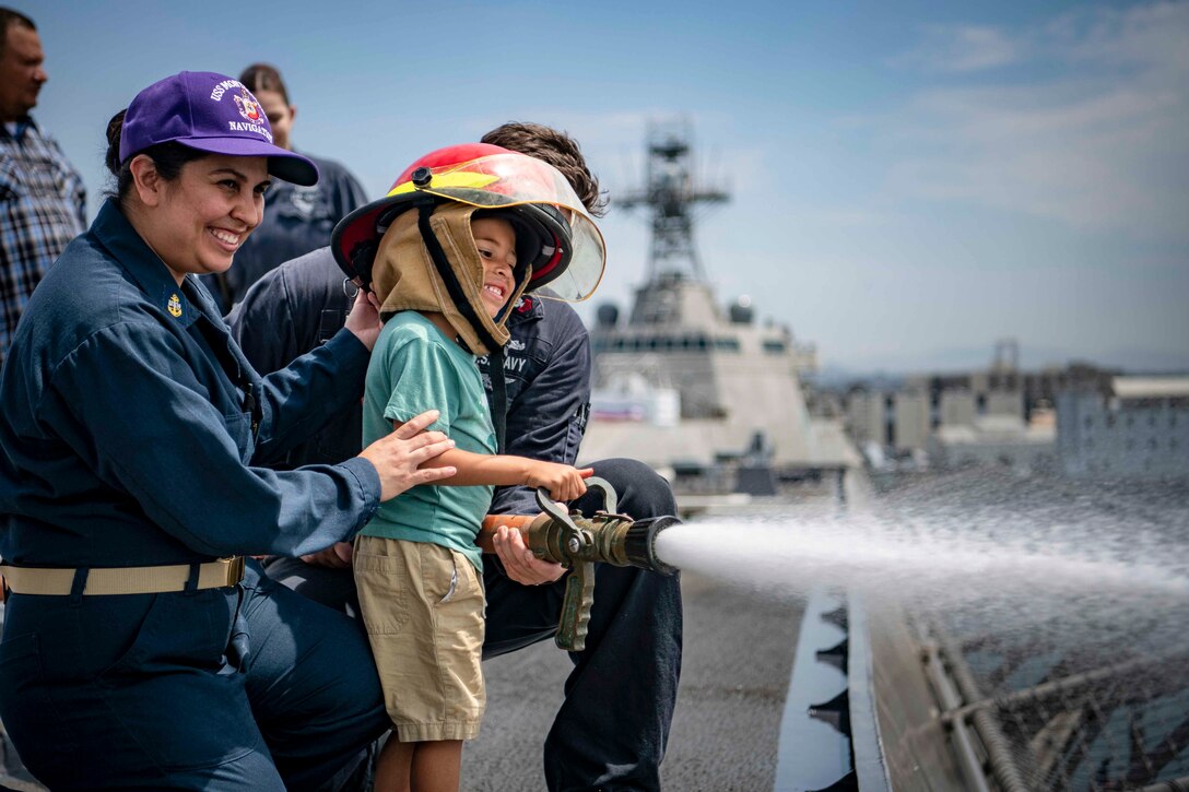 Two sailors assist a child in a helmet hold onto a fire hose on a ship.