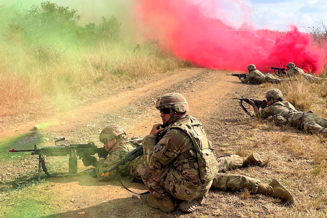 A guardsman kneels next to four guardsmen lying on the ground as green and pink smoke surrounds.