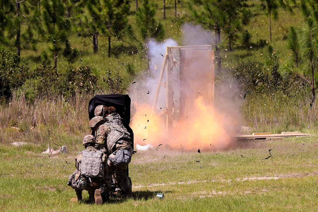 Soldiers stand behind a blast blanket as a door explodes in a field.