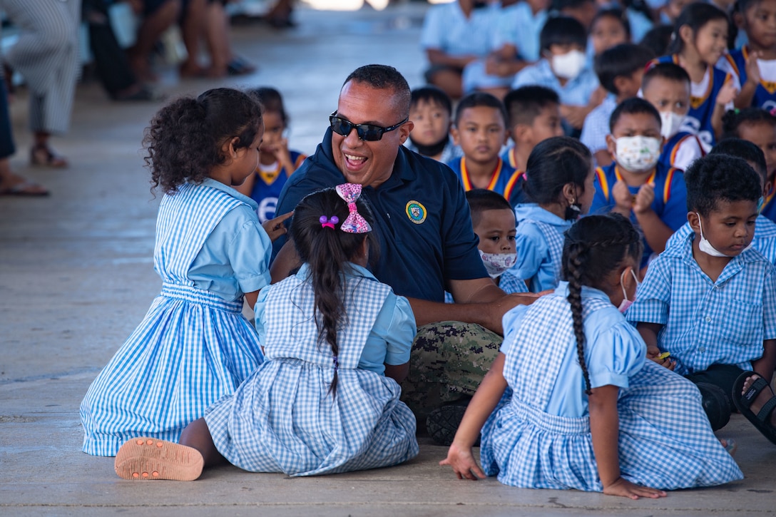 A sailor sits on the floor and is surrounded by children.