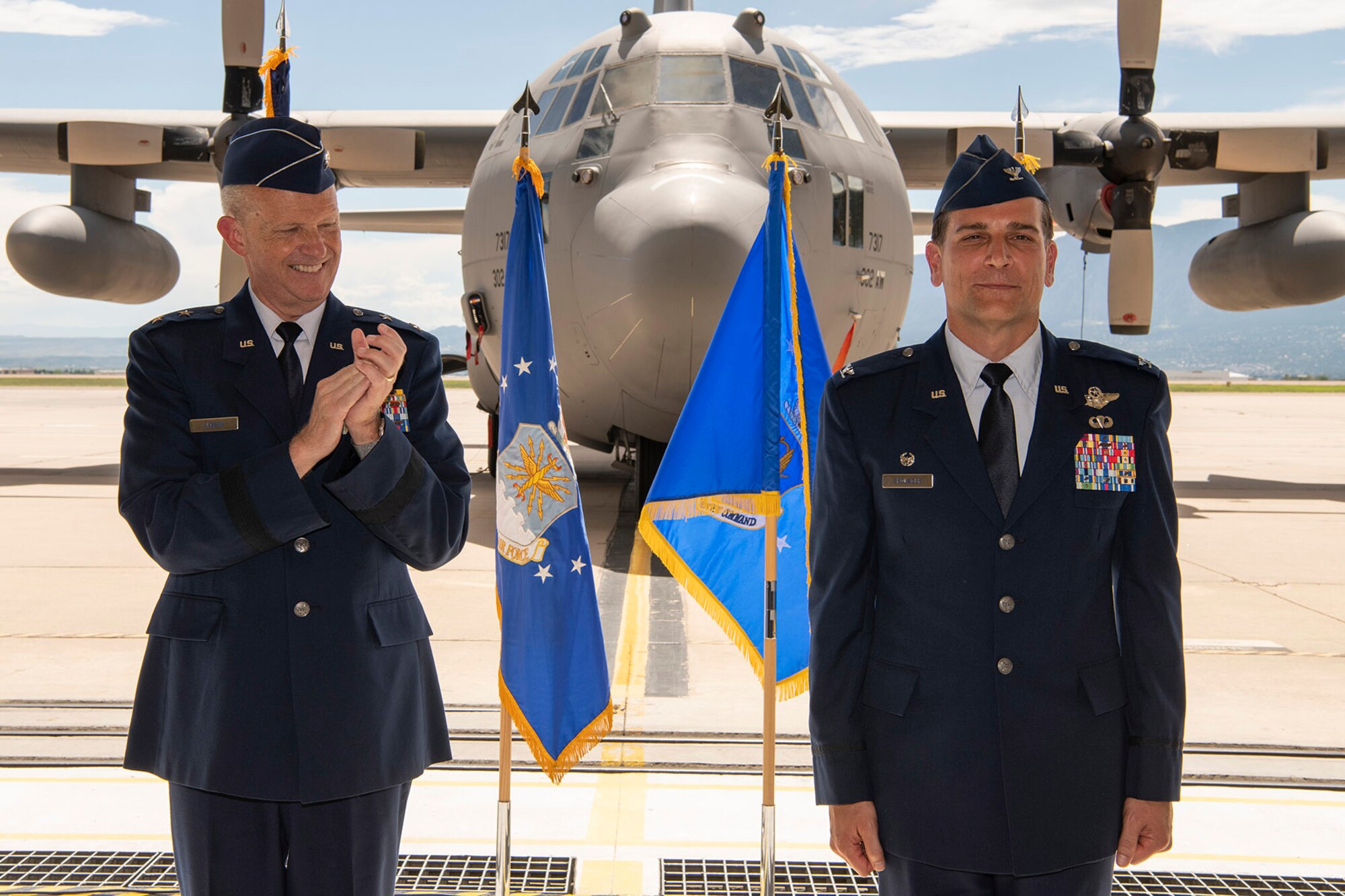 A man in Air Force service dress applauds another man in Air Force service dress with a C-130 in the background.