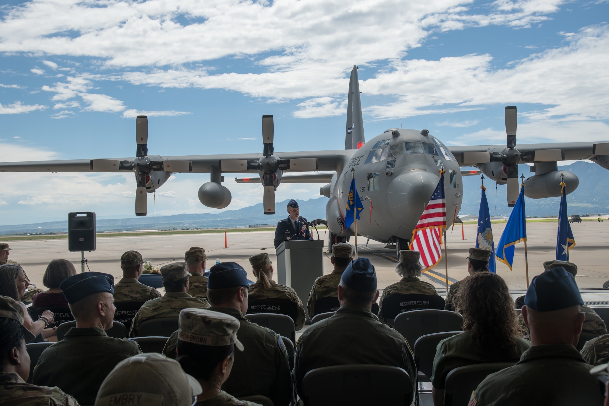 A man in Air Force service dress addresses a small crowd from a podium with a C-130 in the background.