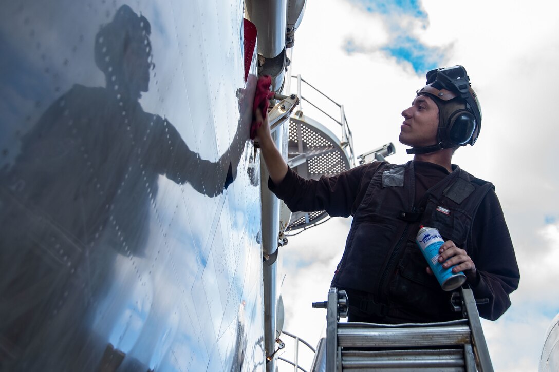 A sailor stands on a ladder and wipes the surface of an aircraft.