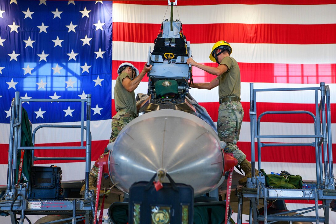 Two airmen standing on ladders install a seat into an aircraft in a hangar.