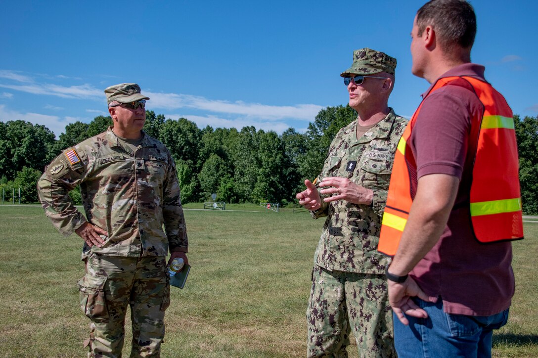 An Army officer, a Navy officer, and a civilian talk in a field.