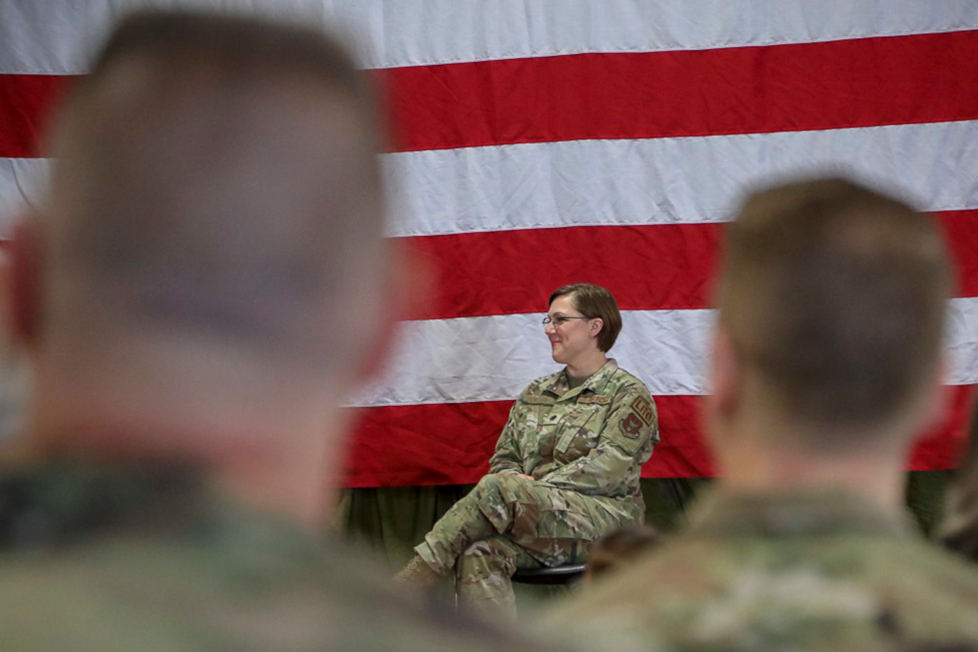 Lt. Col. Tara Horton smiles as she sits in operational camouflaged pattern uniform with the American flag draped behind her.