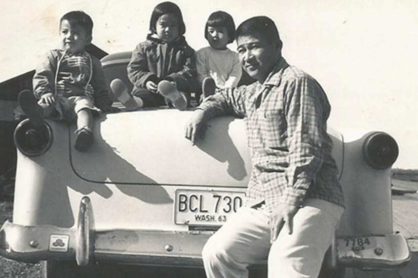 A man sits on a car bumper. Three small children sit on the car’s trunk.