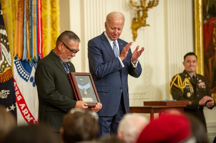 A man looks at a plaque in his hands in front of a crowd as another man claps.