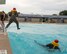 A man is in mid-air over a pool after jumping in, while another man looks on from inside the pool.