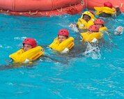 Five aircrew members swim backwards towards a large life raft in a chain formation.