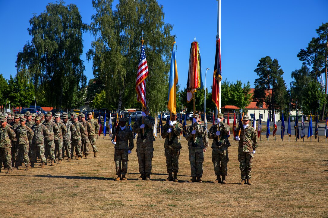 A row of soldiers holding flags stands in formation on a field while others look on.