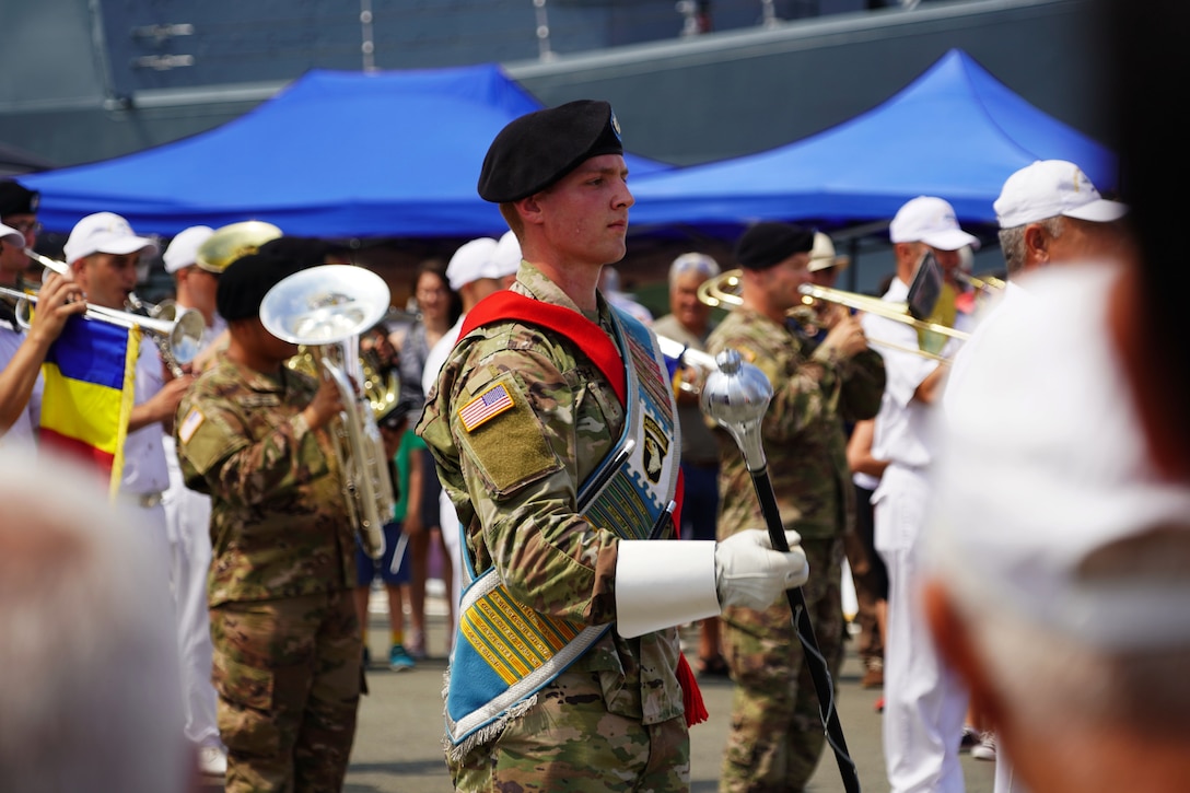 A U.S. soldier holding a baton leads a band along a street with tents in the background.