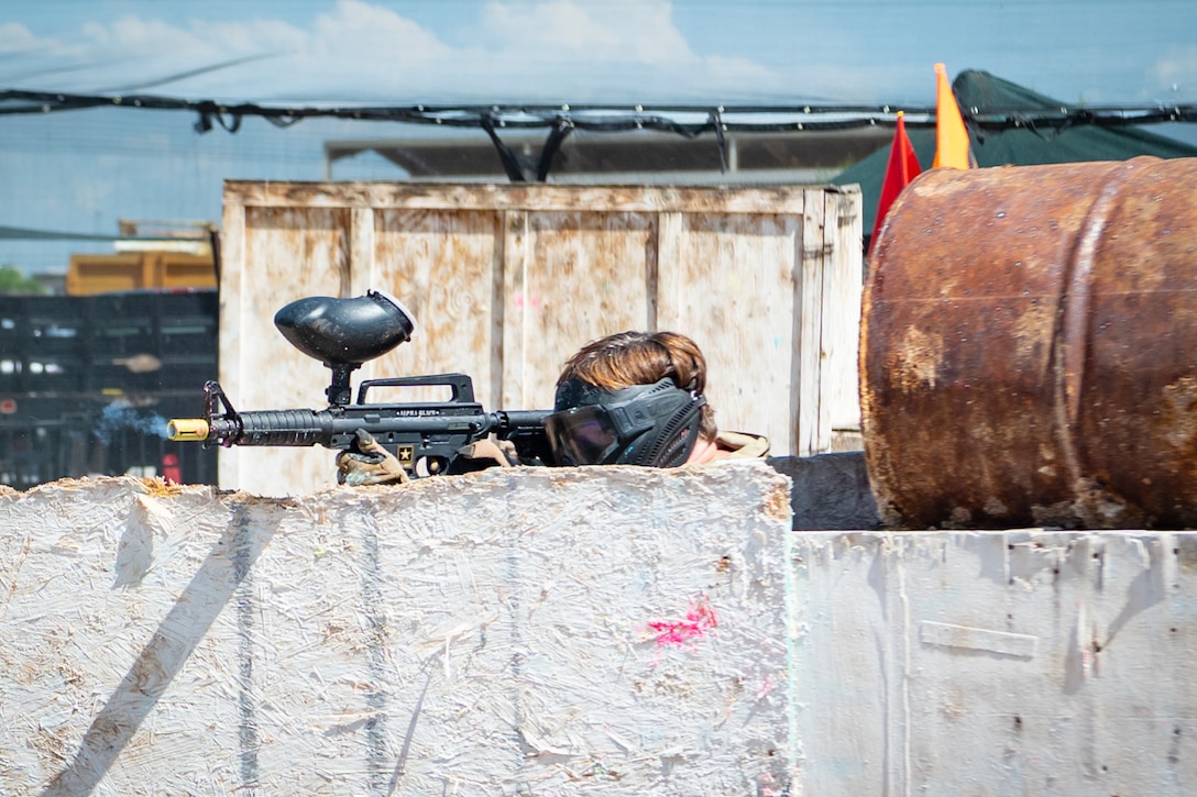 An airman aims a paintball rifle from behind a wall.