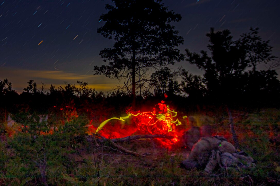 Soldiers move through a wooded area at night as red and yellow lights shine in front of them.